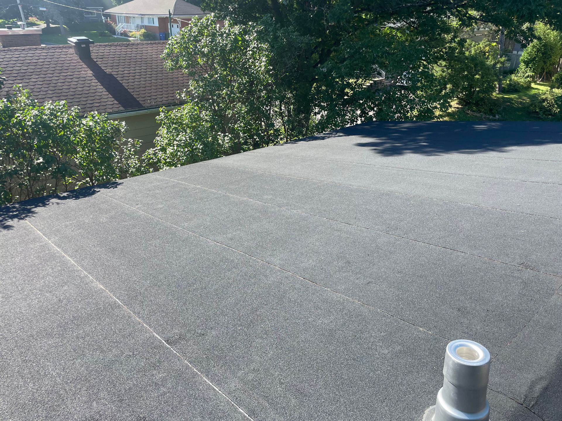 Toit plat gris avec un conduit de ventilation, donnant sur les arbres et les maisons voisines sous un soleil éclatant