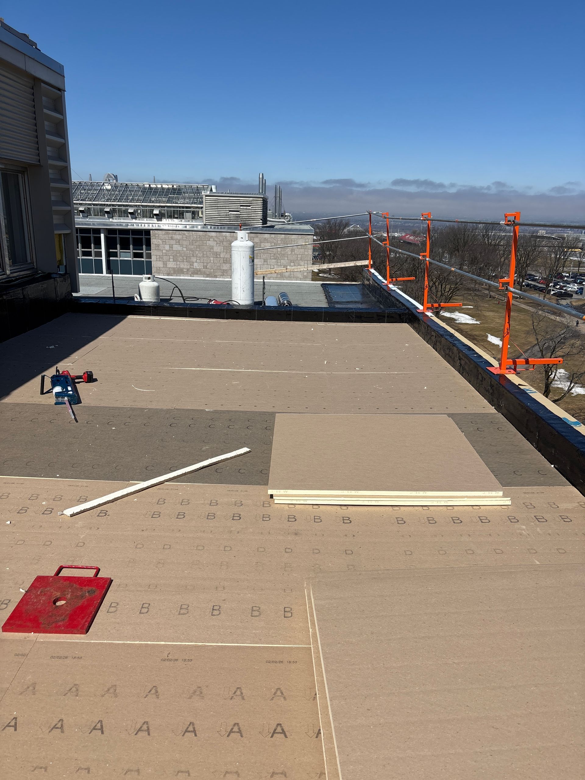Chantier de construction sur un toit avec toiture beige, rambardes de sécurité orange et bâtiments de la ville en arrière-plan