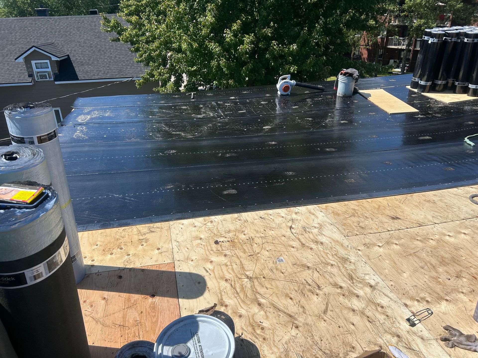 Toiture en construction avec membrane d'étanchéité noire, platelage en contreplaqué, arbres et maisons en arrière-plan