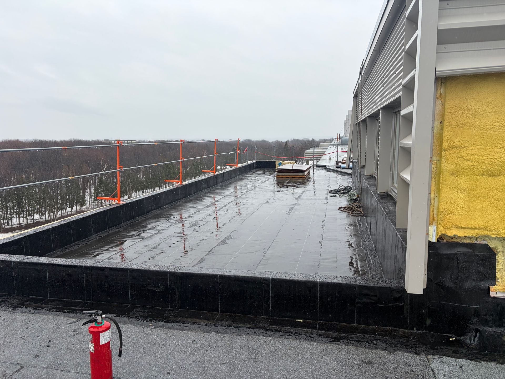 Passerelle sur le toit enneigée, rambarde noire, mur jaune du bâtiment et extincteur au premier plan