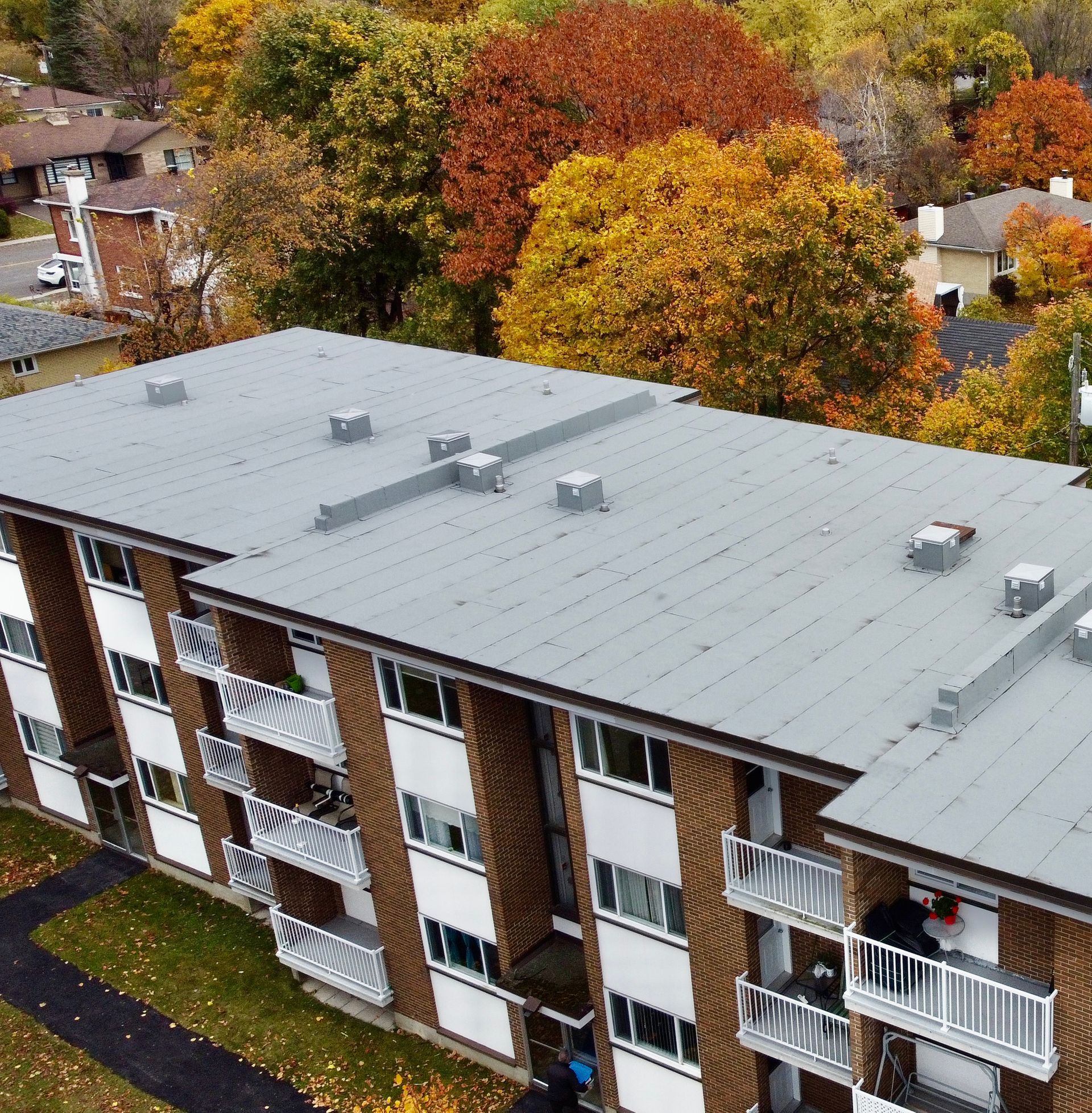 Vue aérienne d'un immeuble d'appartements en briques aux balcons blancs, bordé d'arbres d'automne au feuillage coloré.