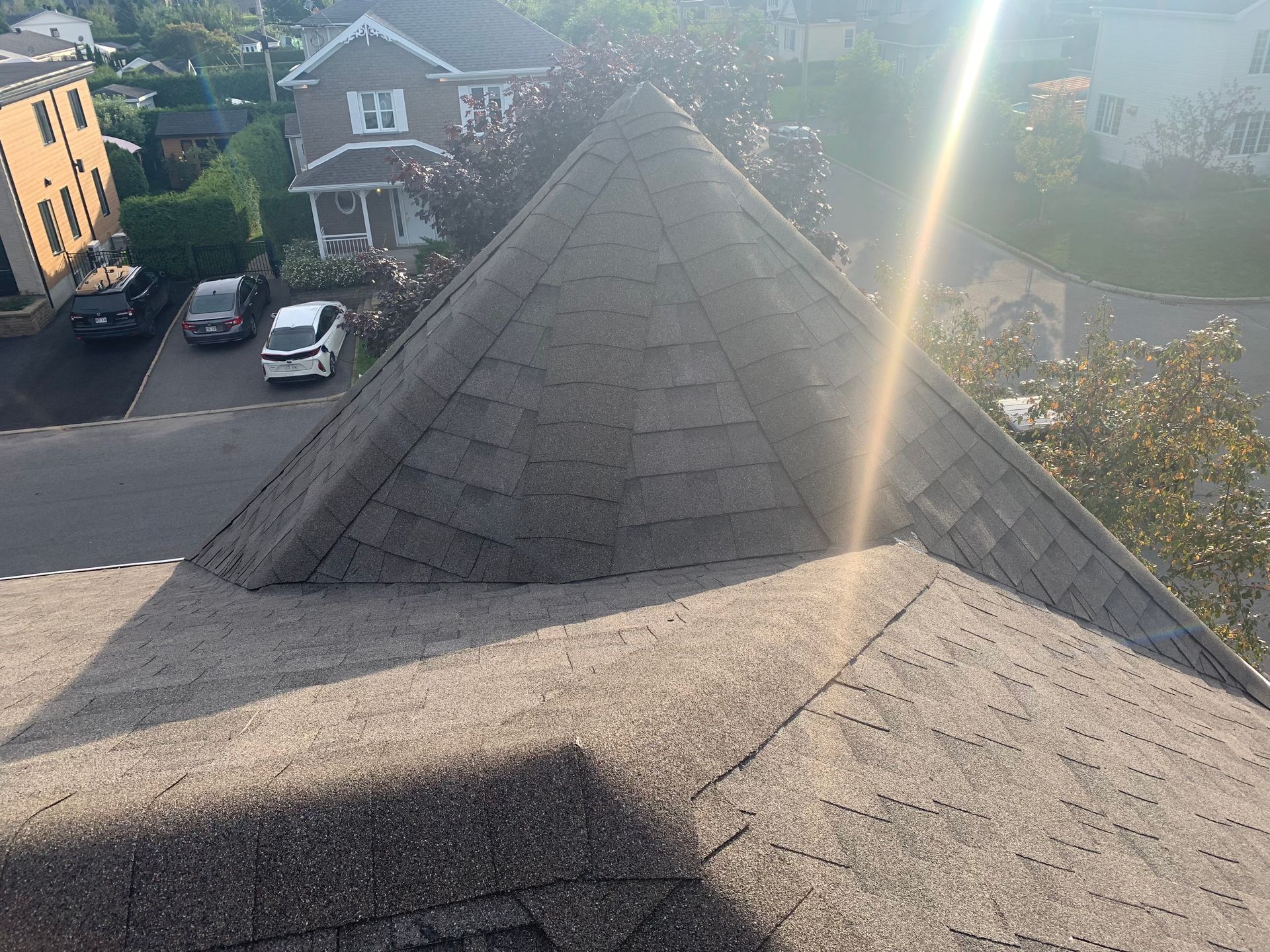 Vue depuis un toit de bardeaux donnant sur une rue résidentielle, avec les reflets du soleil et les maisons voisines.