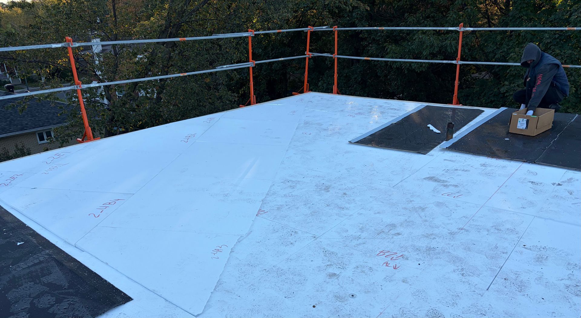 Toiture en construction avec étanchéité blanche, rambardes de sécurité et un ouvrier agenouillé près d'outils