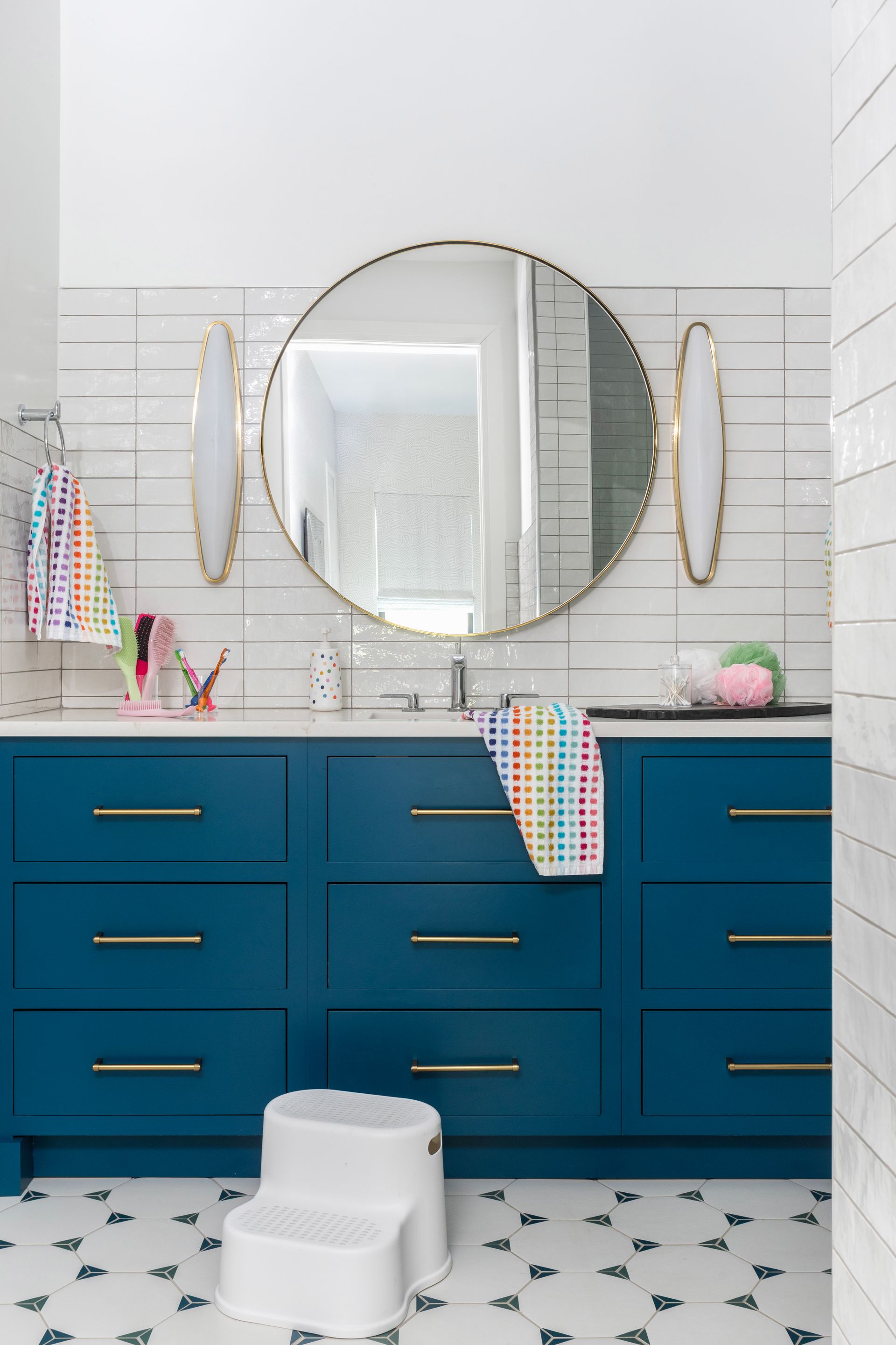 Blue bathroom vanity with gold accents, round mirror, white step stool, and patterned tile floor.
