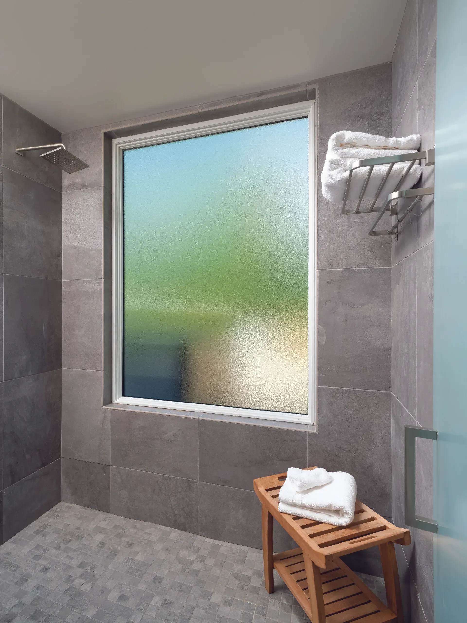 Bathroom with gray tiled walls, shower head, window with frosted glass, towels on a rack and bench.