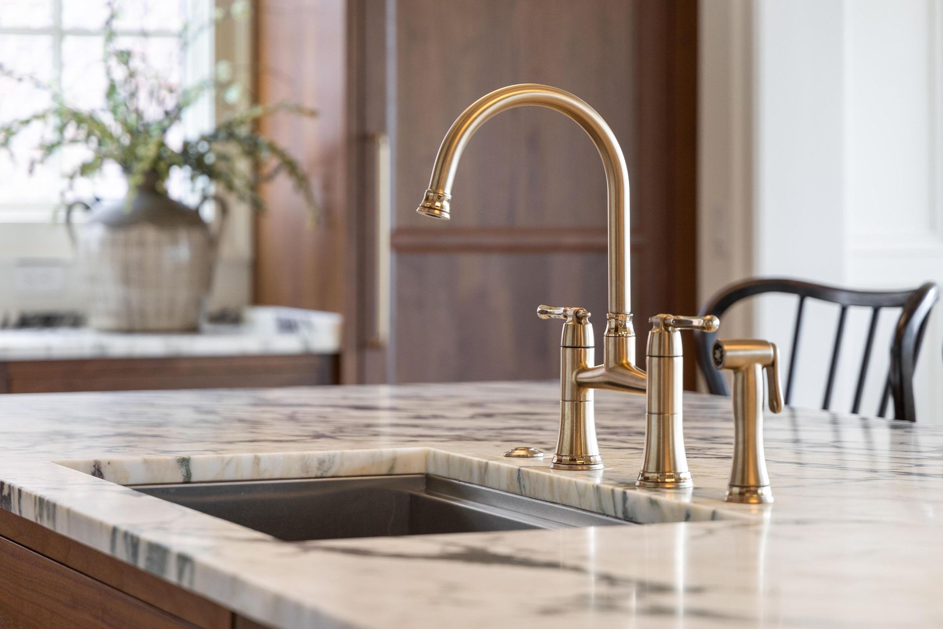 Gold kitchen faucet on a marble countertop with a stainless steel sink and a window in the background.