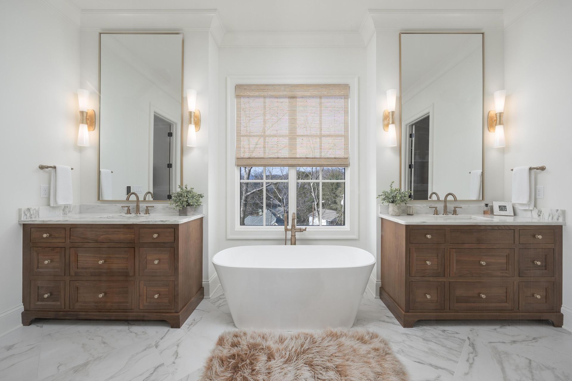 Spacious white bathroom with two wooden vanities, large mirrors, a freestanding tub, and a window with a blind.