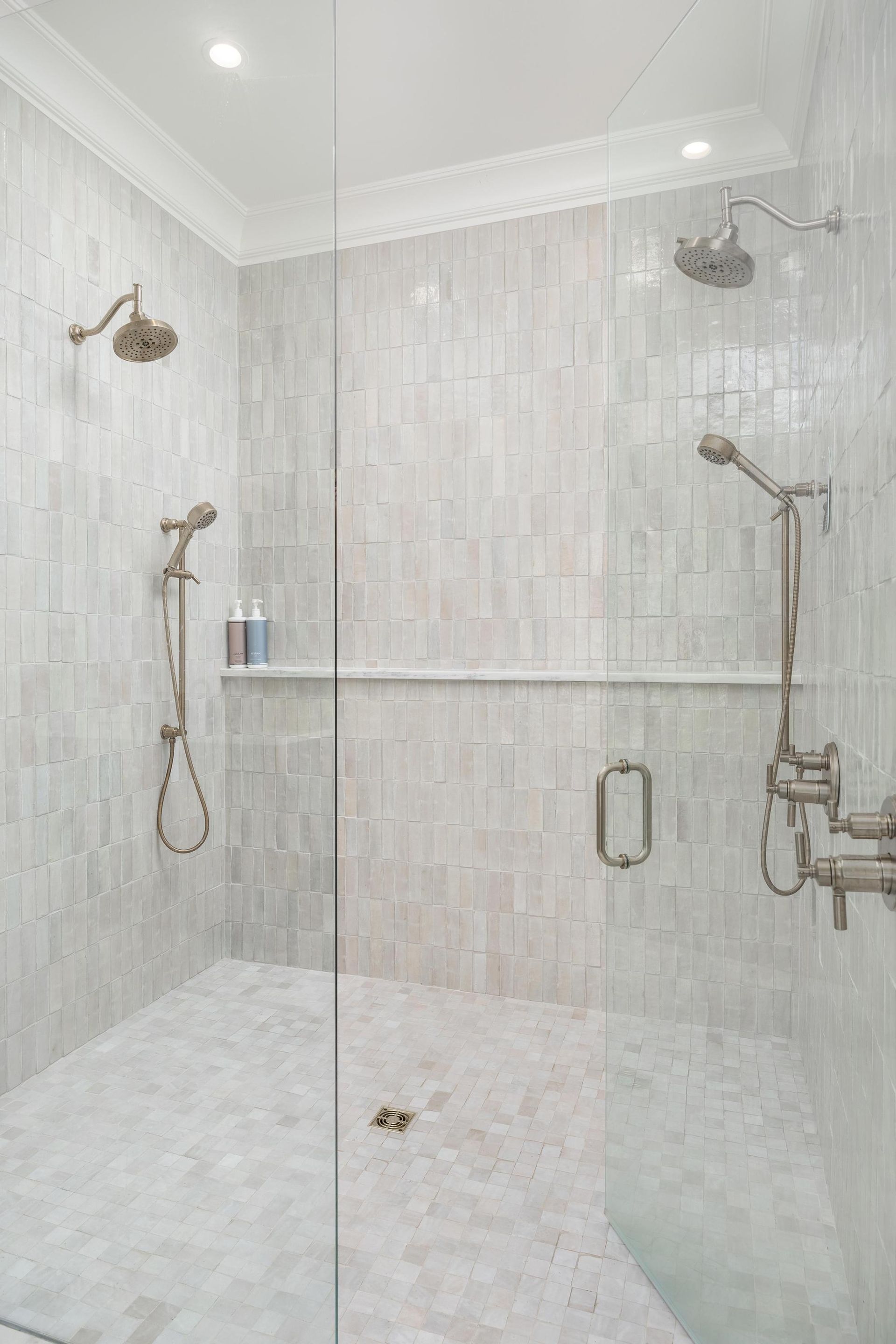 Tiled shower with three showerheads, glass door, and pebble floor. Off-white with bronze fixtures.