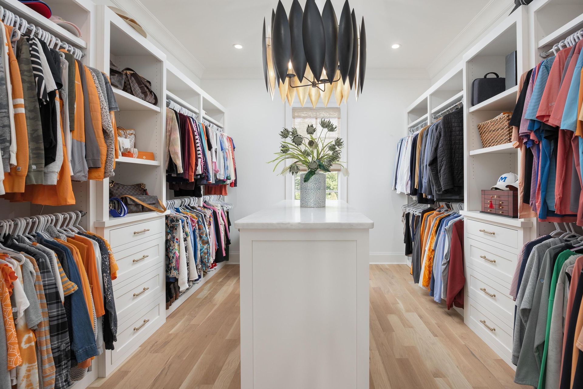 Walk-in closet with white shelves, drawers, and rows of clothing. Light fixture hangs overhead.