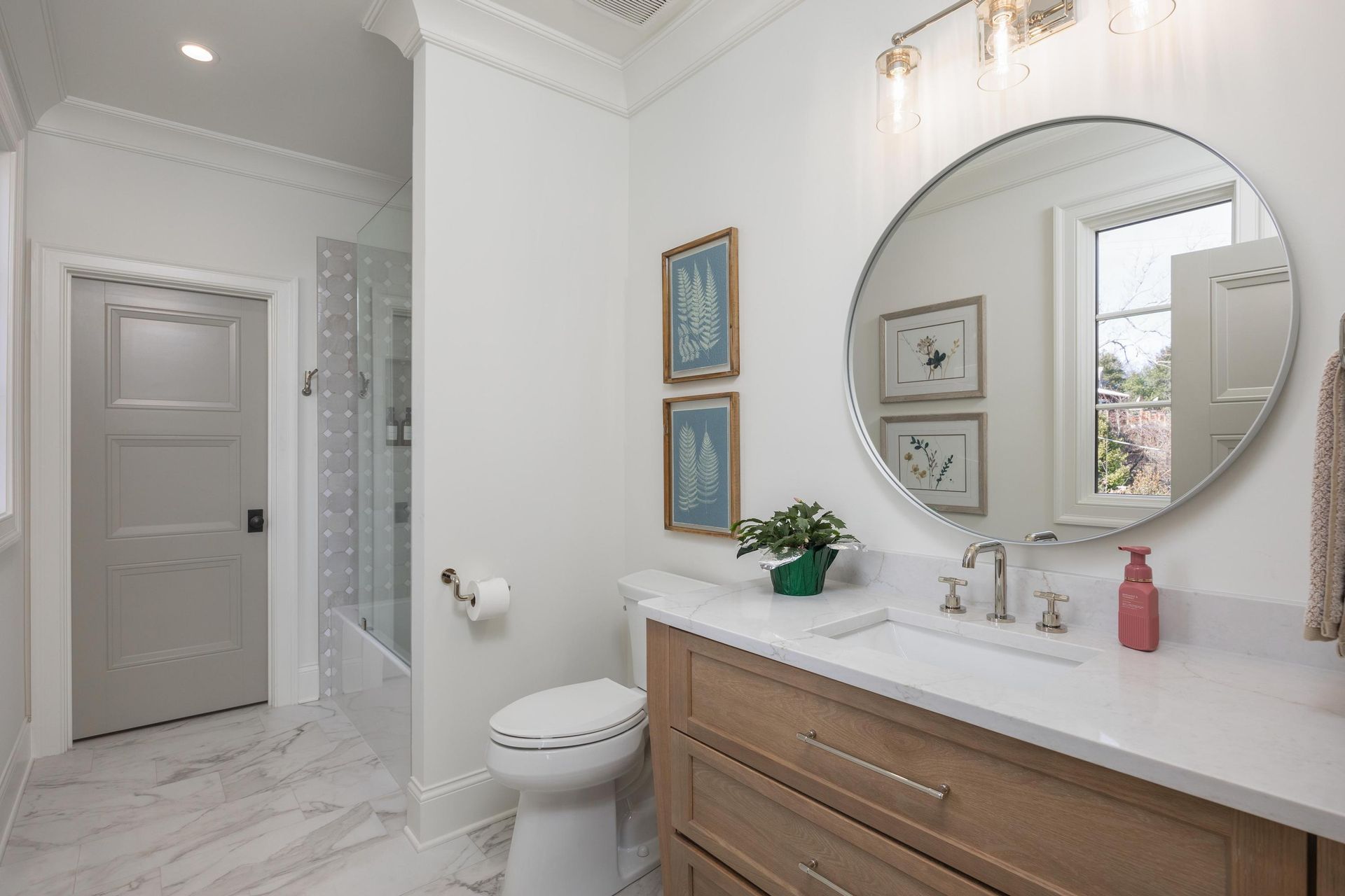 Bathroom with light wood vanity, white countertop, round mirror, and framed botanical prints.