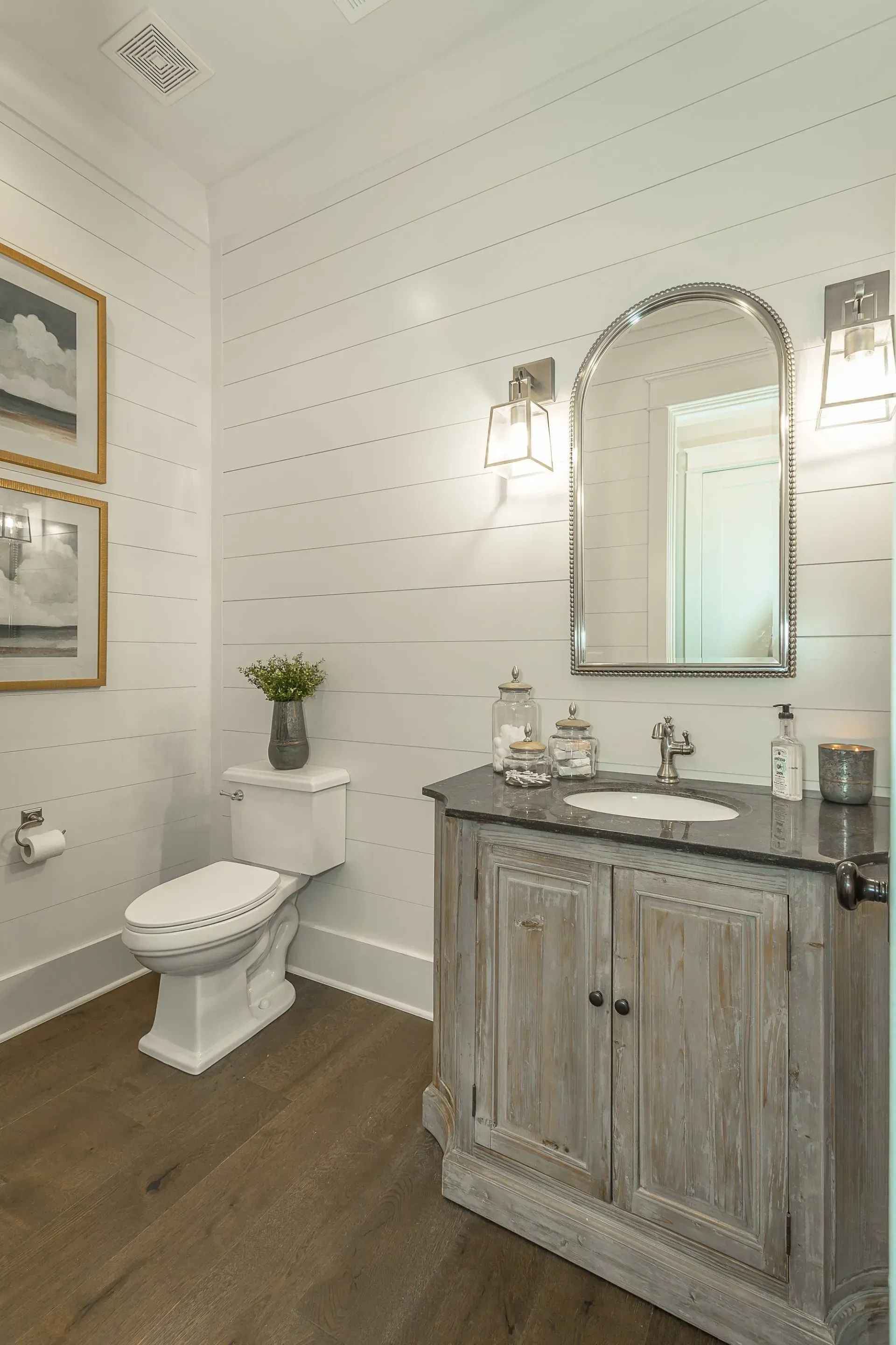 Bathroom with white shiplap walls, wooden vanity with dark countertop, arched mirror, and hardwood floor.