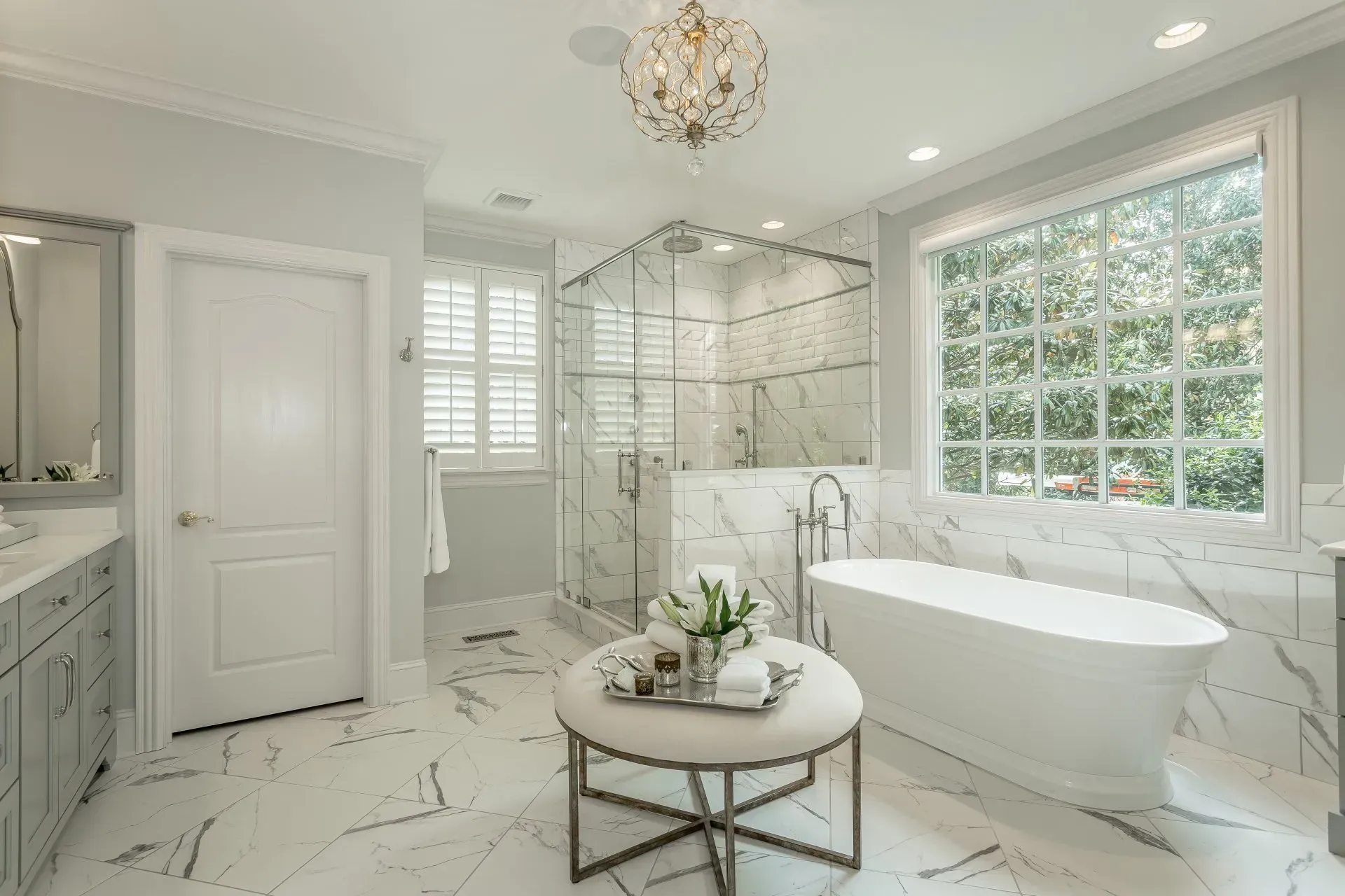 Elegant white marble bathroom with a freestanding tub, glass shower, and round ottoman.