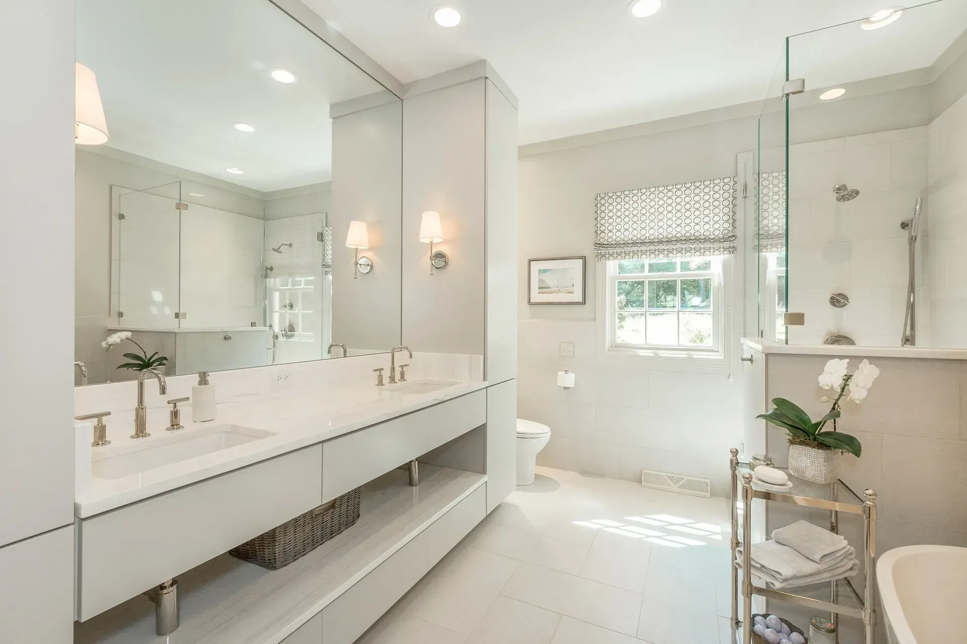 Spacious, modern bathroom with white tile and fixtures, large mirror, and shower, window with patterned shade.