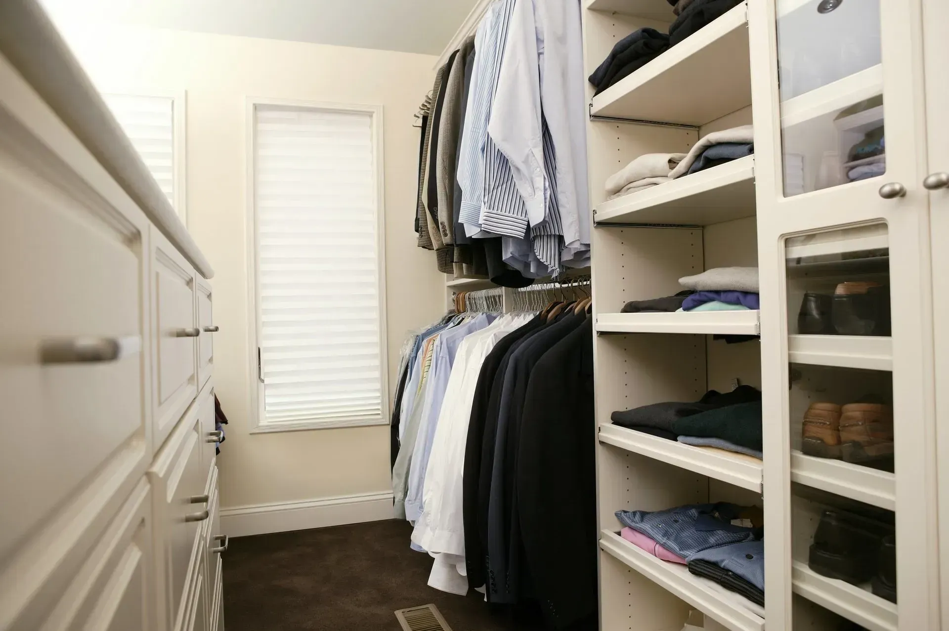 Walk-in closet with hanging clothes, shelves, and drawers, cream-colored cabinetry, brown floor, window with blinds.