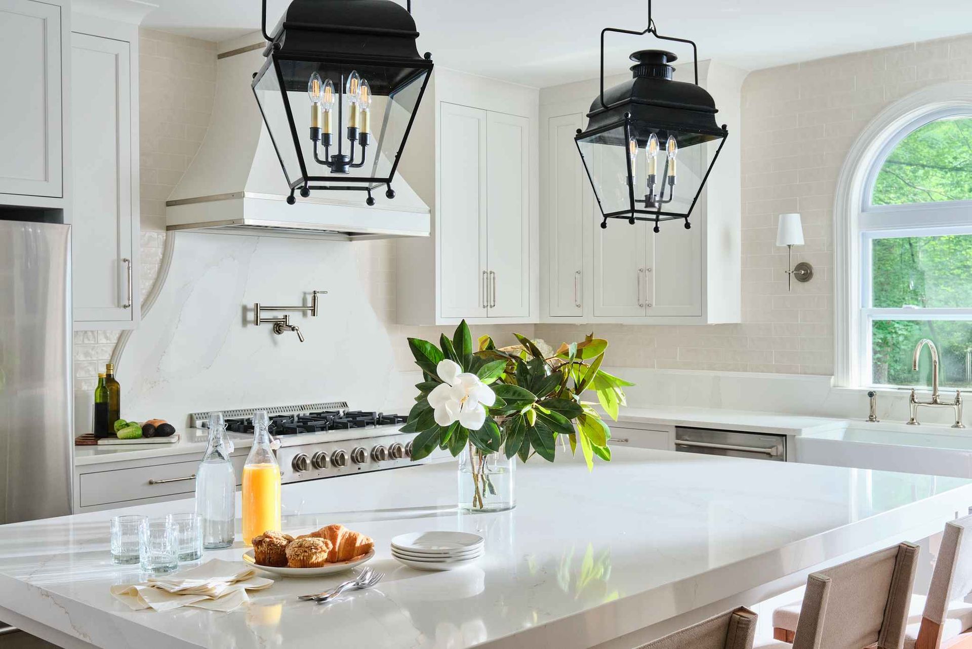 Bright white kitchen with a large island. Black lanterns hang overhead. Fresh flowers and breakfast set out.