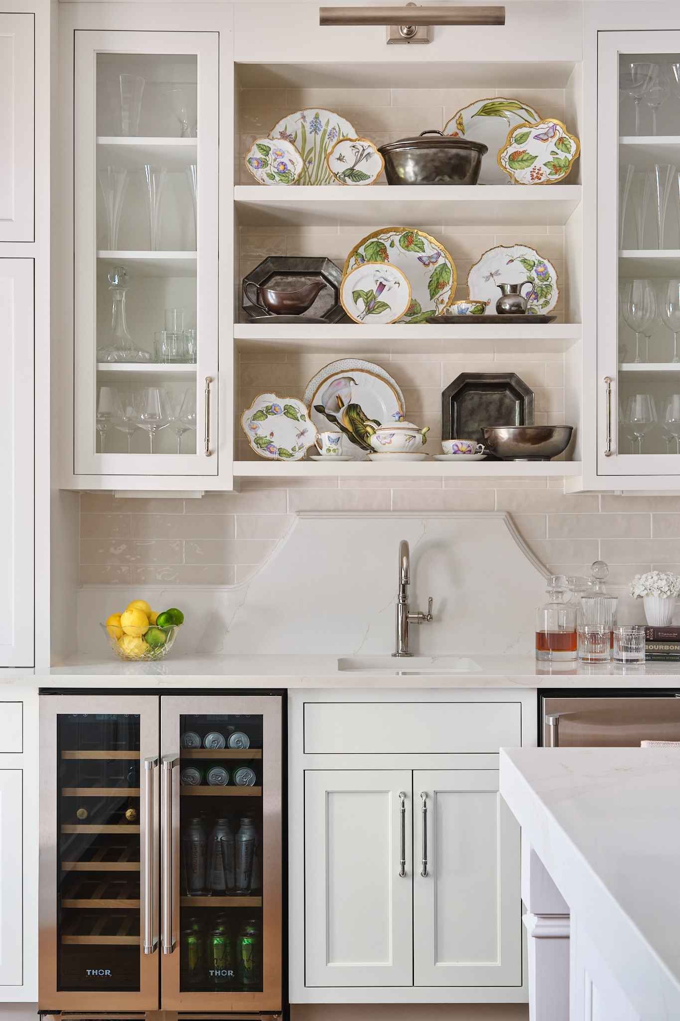 Built-in kitchen shelving with display plates and glasses. Wine cooler below sink, white cabinets.
