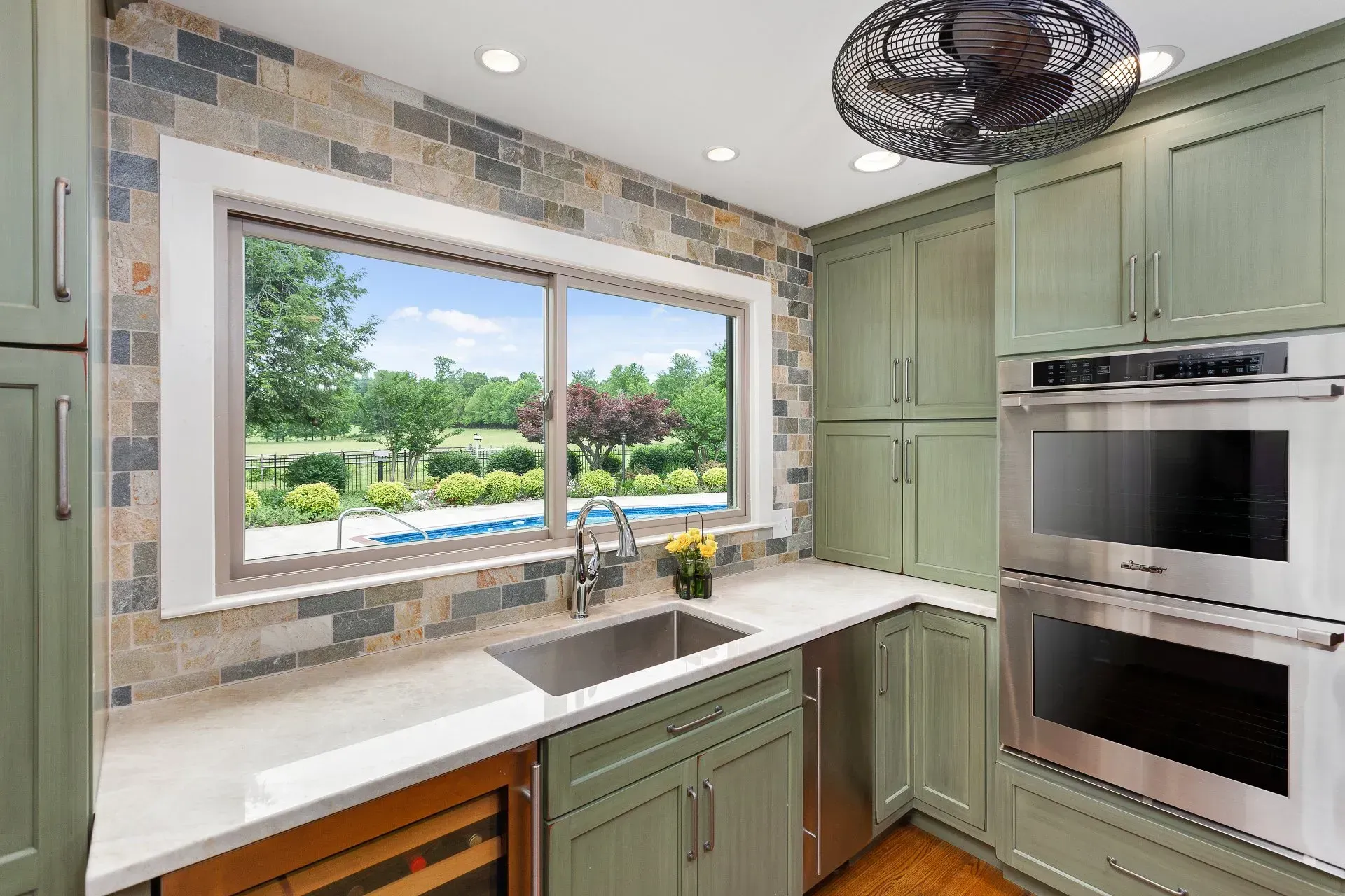 Kitchen with sage green cabinets, stone backsplash, stainless steel appliances, and a window overlooking a pool.
