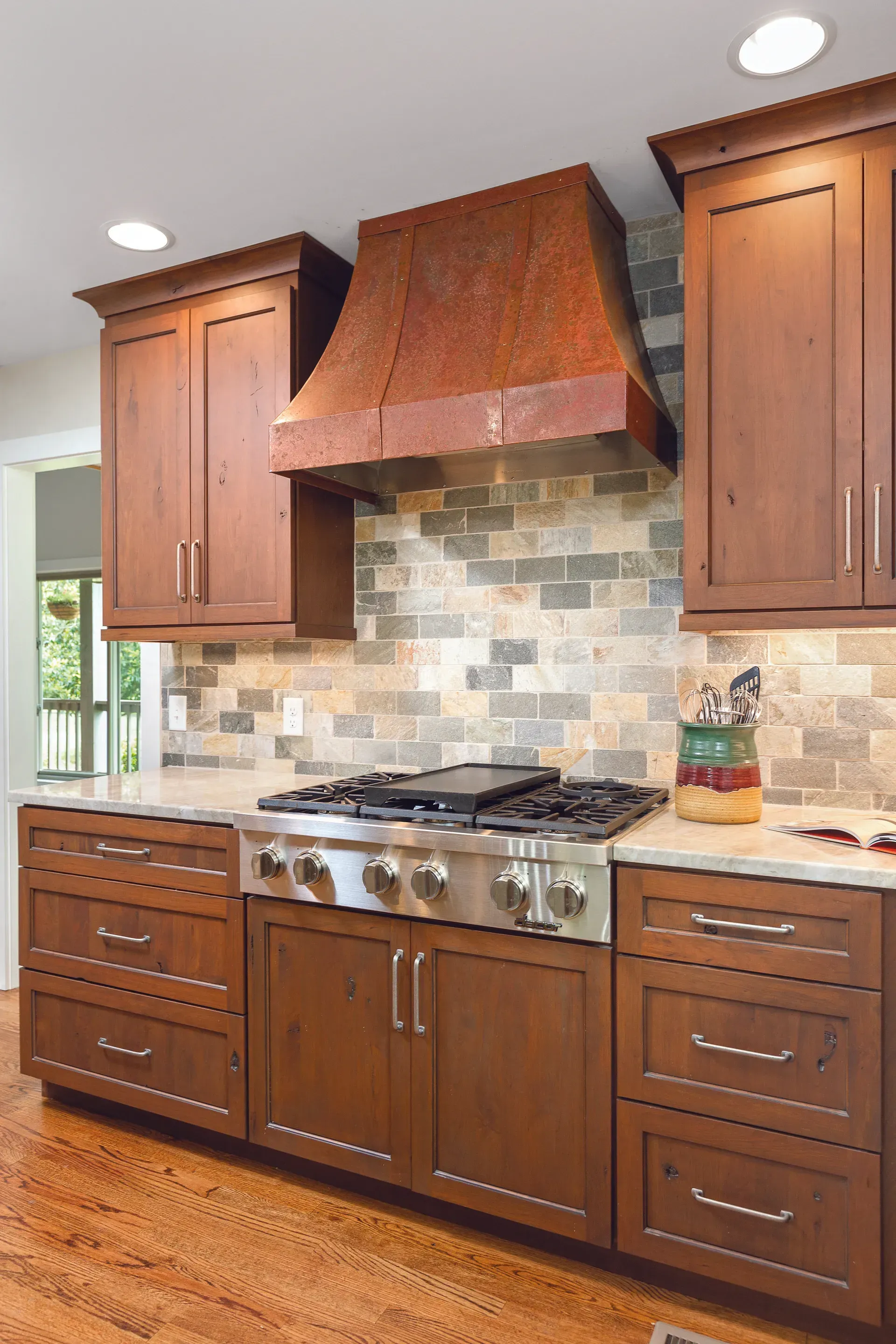 Kitchen with stainless steel stove, copper range hood, brown cabinets, and stone backsplash.