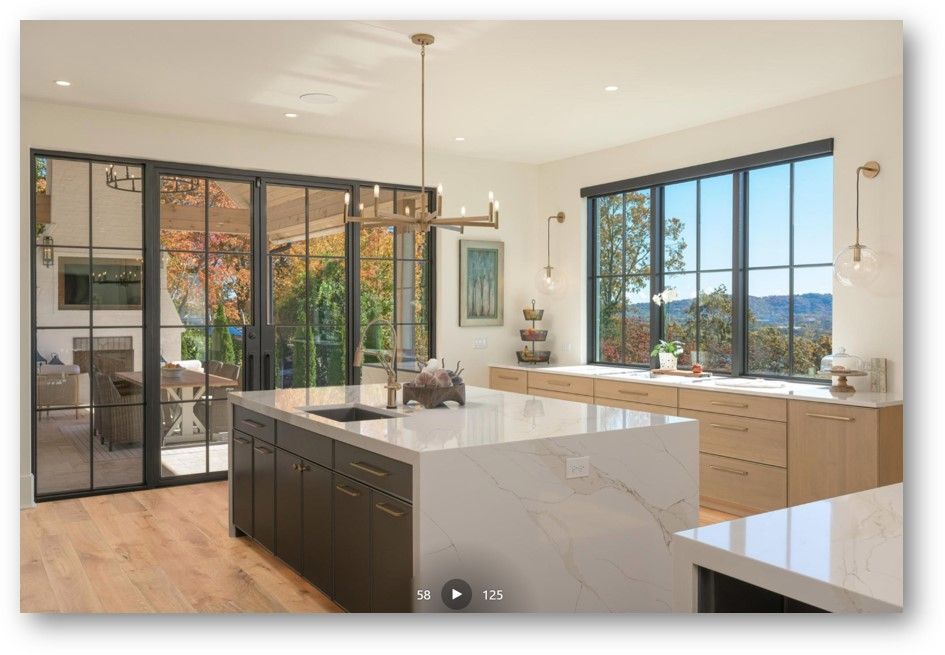 Modern kitchen with large island, black-framed windows/doors, natural light, and outdoor view.