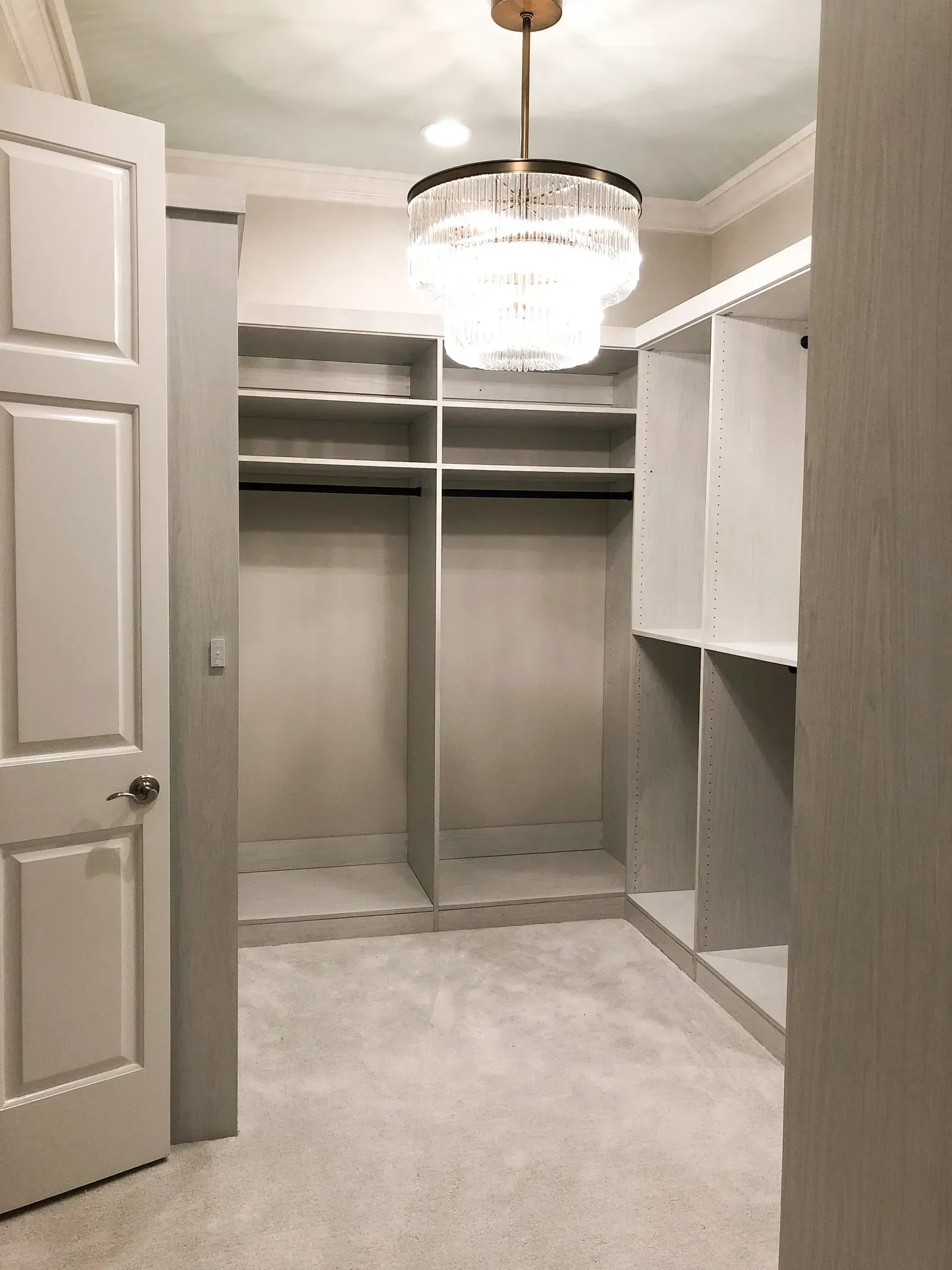 Empty walk-in closet with gray shelves and carpet, ornate chandelier, and white door.