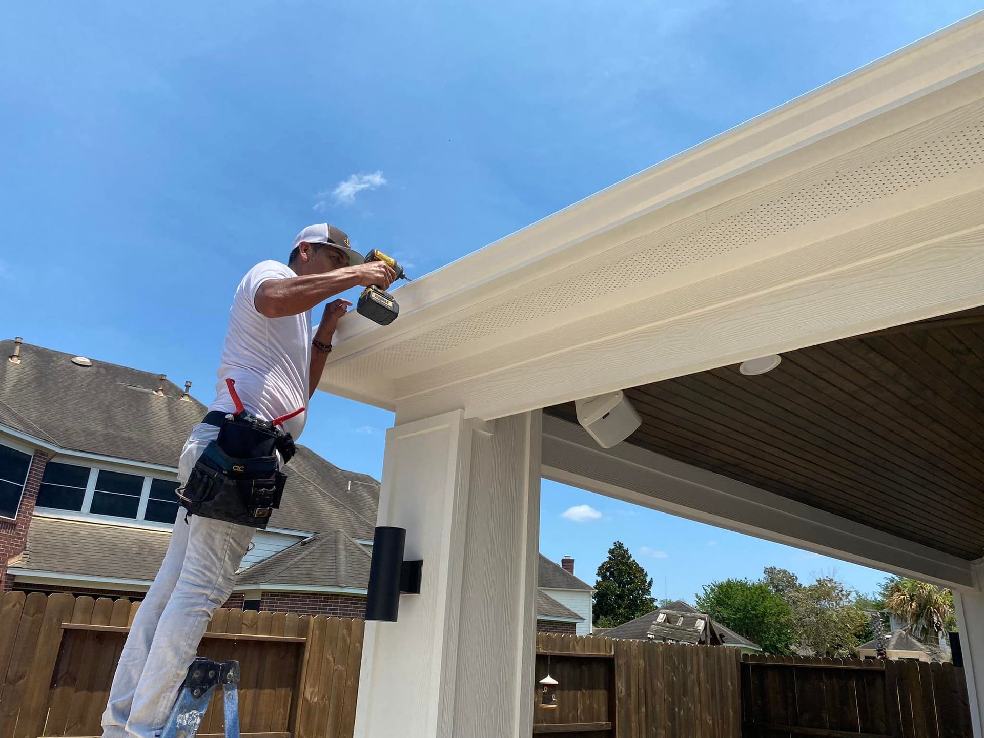 Man on a ladder using a drill to install beige trim on an outdoor structure with brown ceiling.