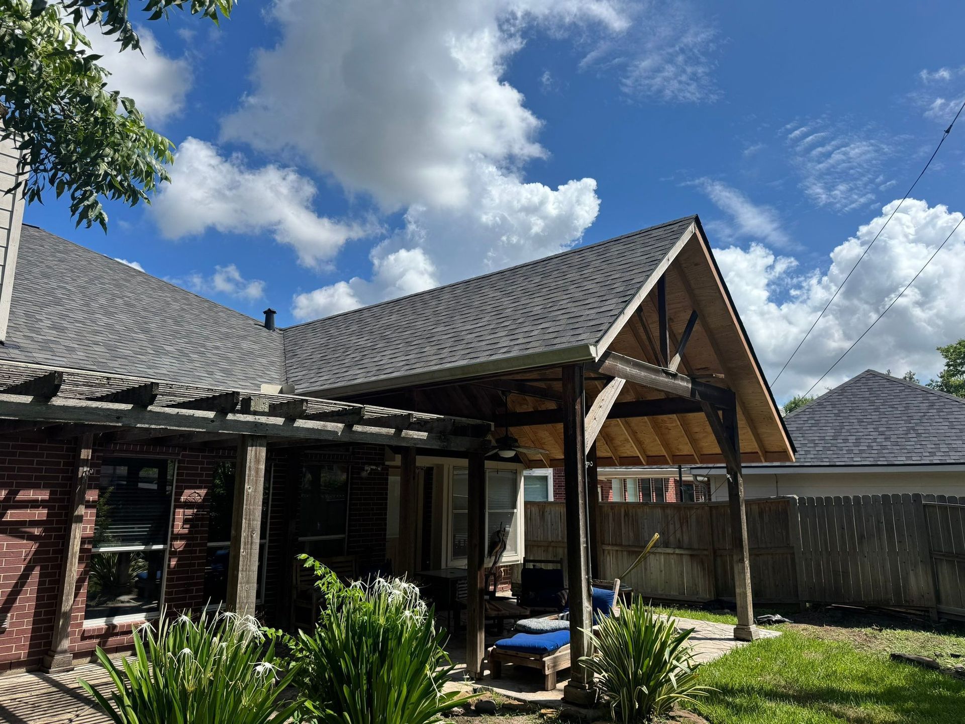 Backyard view of a brick house with a pergola and covered patio, blue sky with fluffy clouds.