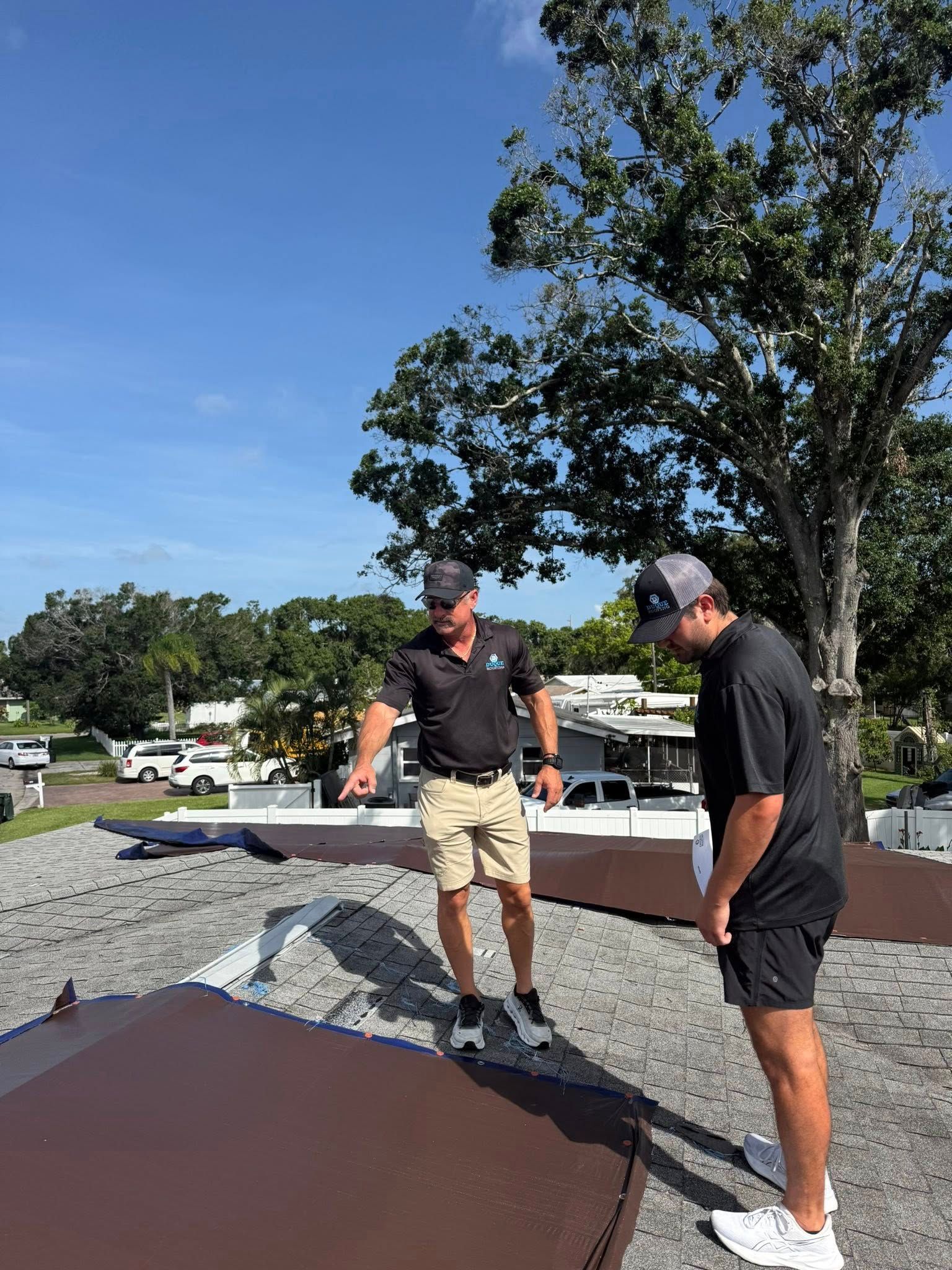 Two men inspecting a roof on a sunny day. One points at a dark brown section. Green tree in background.