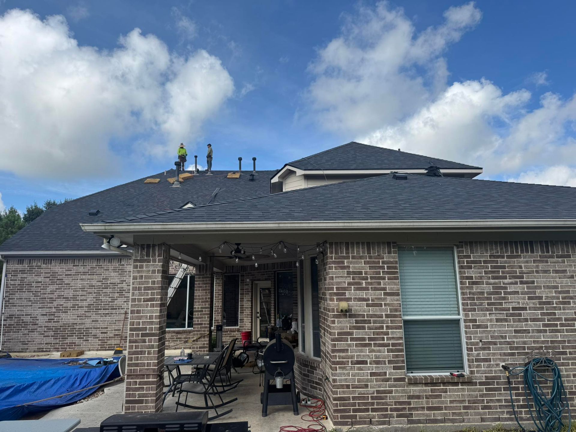 Roofers on a dark shingle roof against a partly cloudy sky. Brick house with patio and pool.