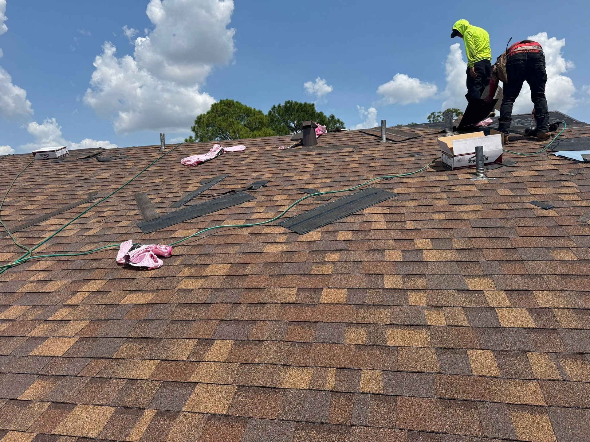 Two roofers installing shingles on a roof under a blue sky with clouds.