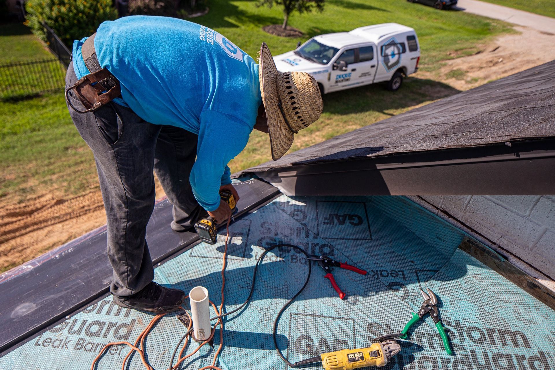 Roofer in blue shirt and straw hat works on a roof. Tools and a work truck are visible.