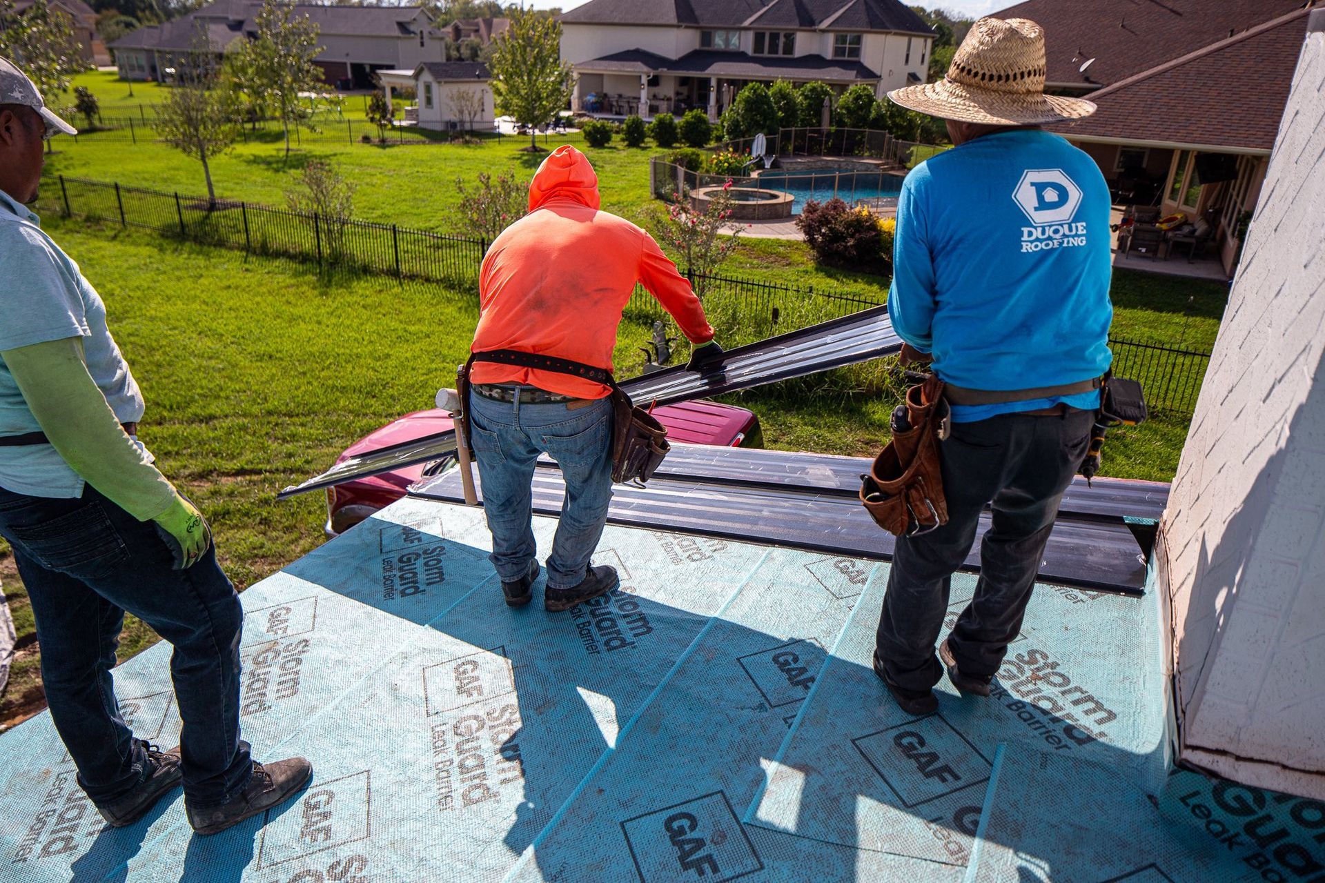 Three roofers working on a roof, sunny day, one in orange hoodie, blue underlayment visible.
