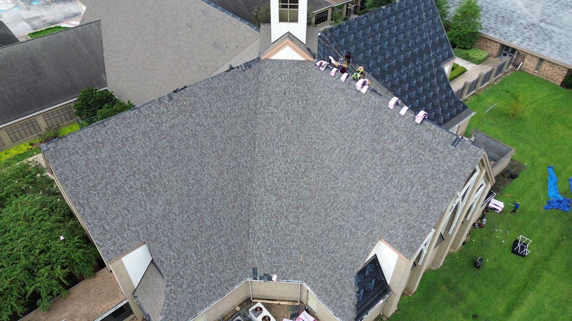 Workers on a gray shingled roof of a house, surrounded by green grass and other buildings.