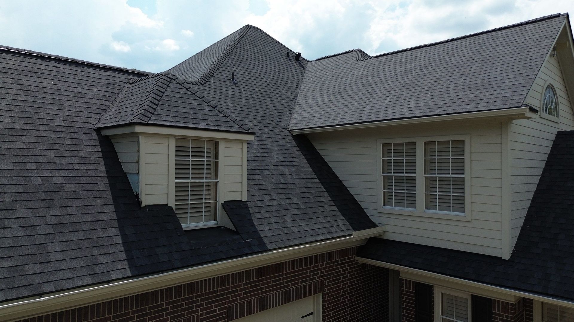 Dark gray shingled roof with dormers, brick facade, and tan trim under a cloudy sky.