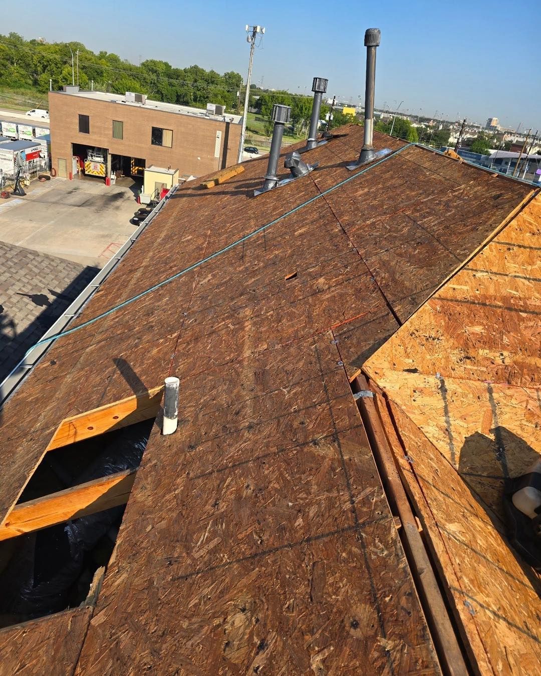 A partially shingled roof with chimneys and exhaust pipes. Buildings and trees in the background under a blue sky.