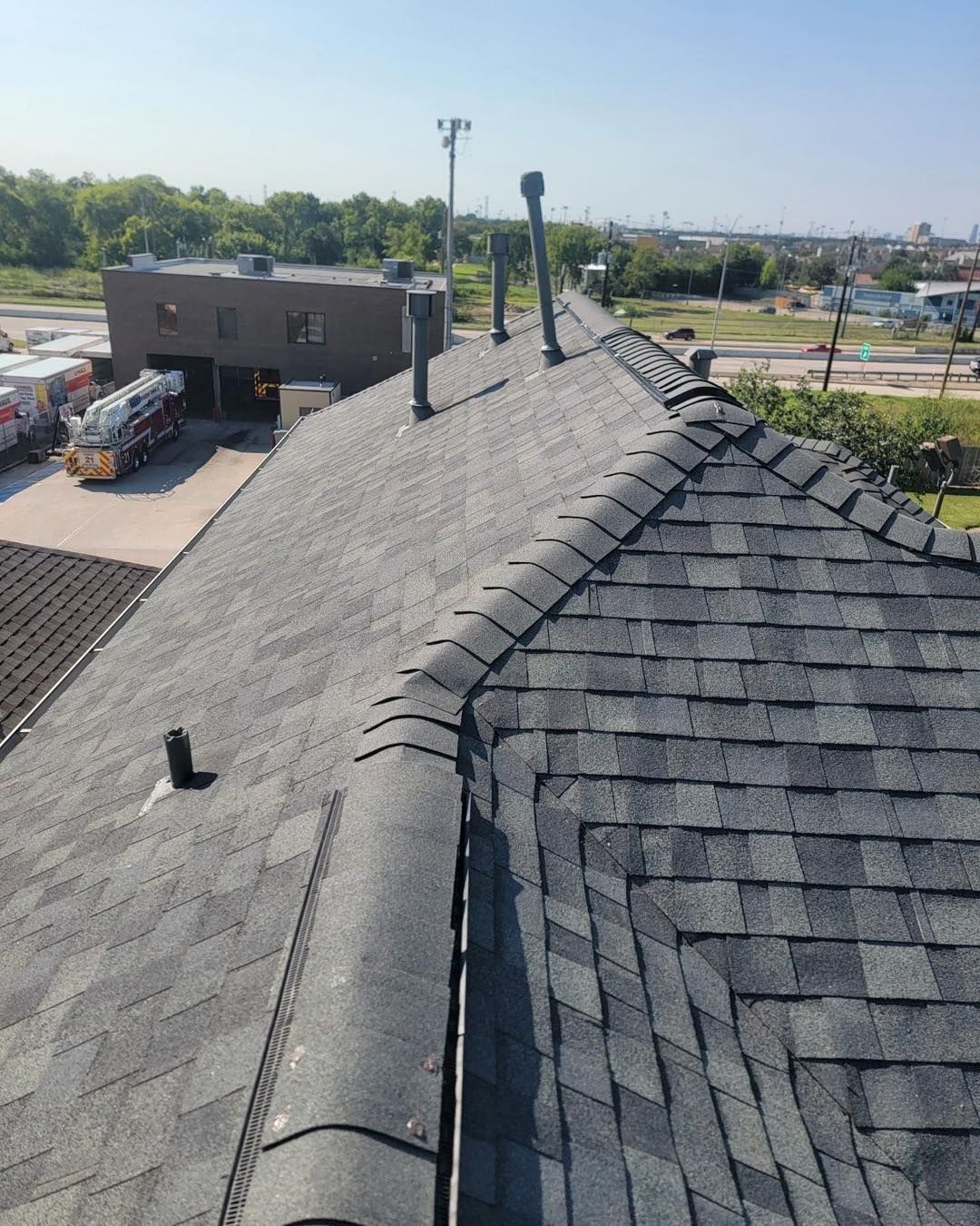 Gray asphalt shingle roof with vents, against a background of a building and industrial area.