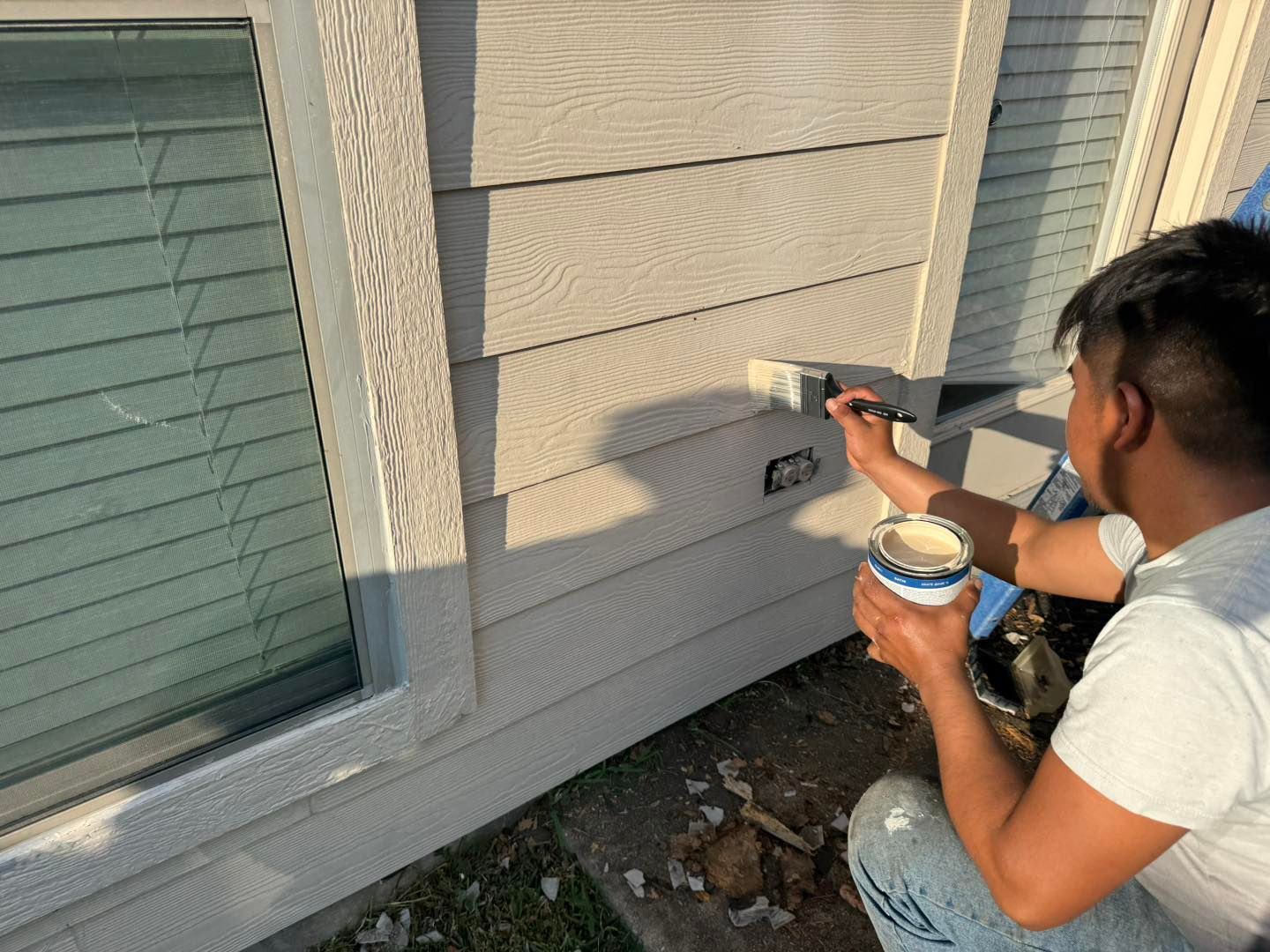 Person painting exterior wall with a brush, near a window. Beige siding.