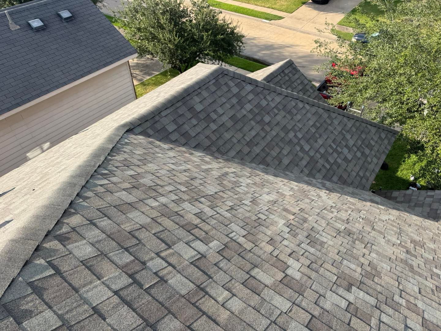Overhead view of a gray shingled roof on a residential building, with trees and a second roof in the background.