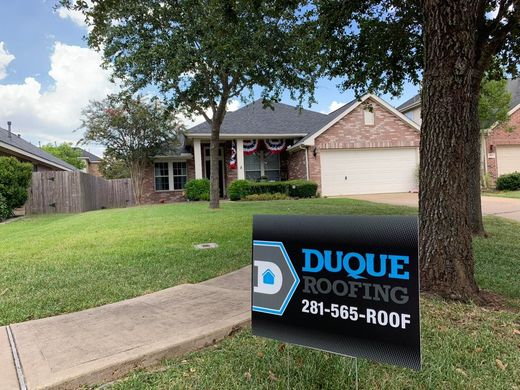 House with a Duque Roofing sign in the yard; green grass, trees, and blue sky.