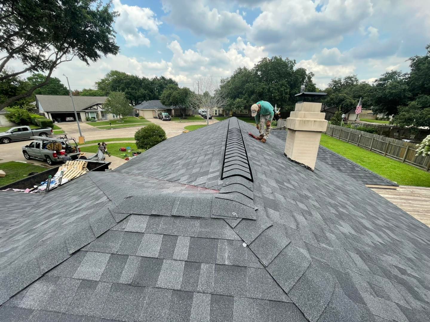 Person working on a gray shingle roof. Ladder and chimney visible, surrounded by trees and houses under a cloudy sky.