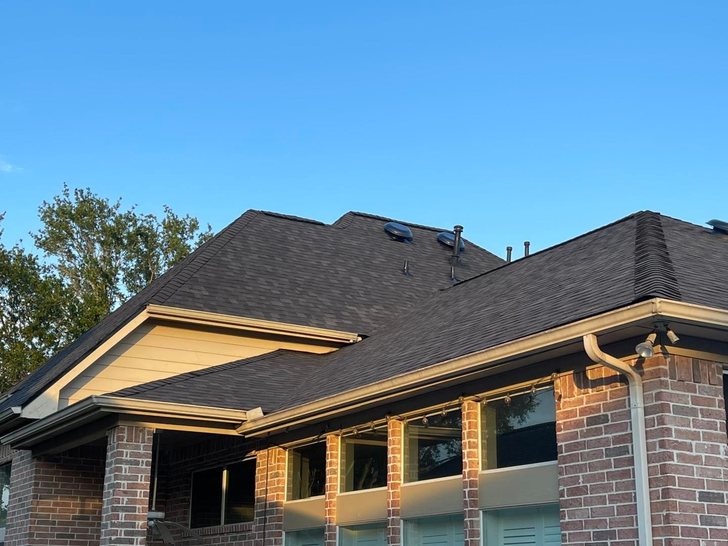 Residential home with dark shingle roof, brick facade, and windows against a blue sky.