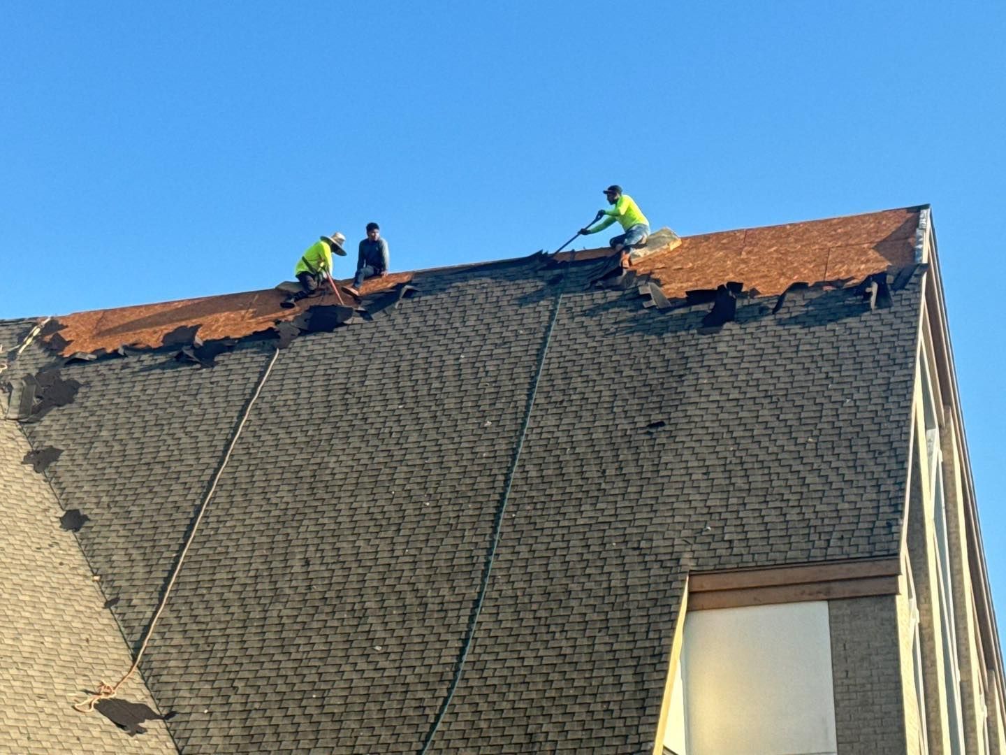 Three workers in safety vests removing old roof shingles on a building under a blue sky.