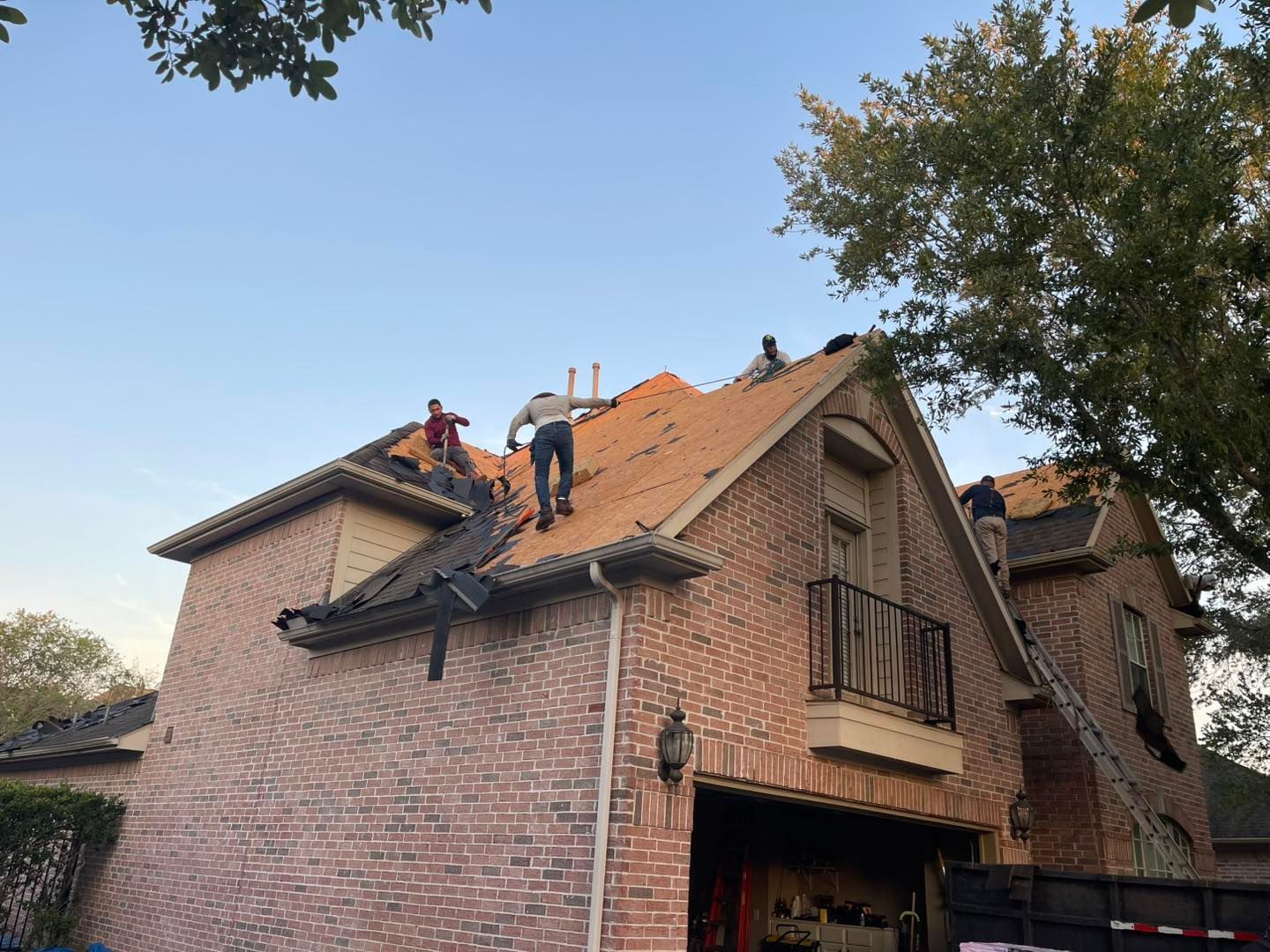 Roofers working on a two-story brick house, removing shingles. A tree and blue sky are in the background.