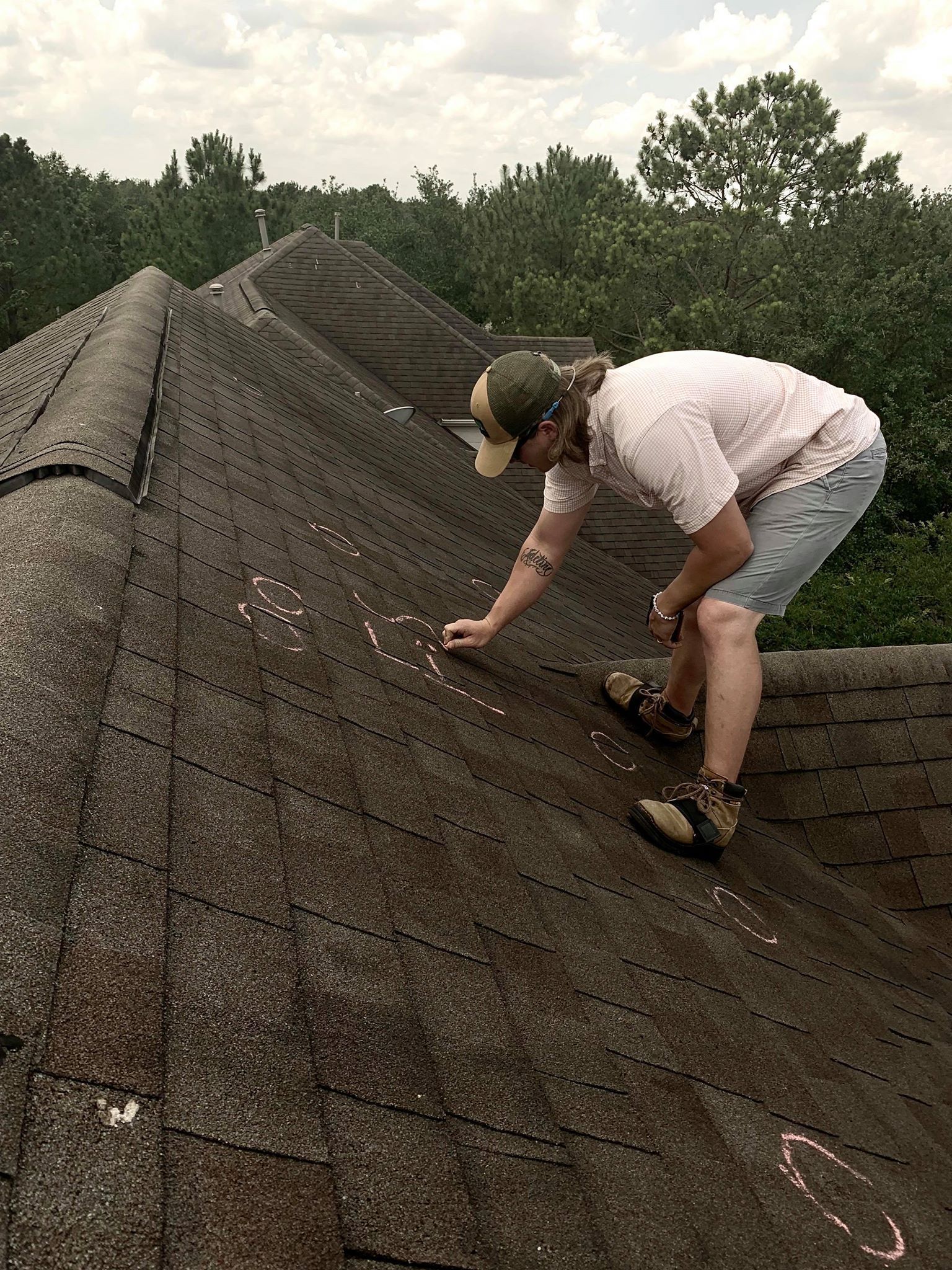 Person on a roof inspecting shingles, trees in the background. Cloudy sky.