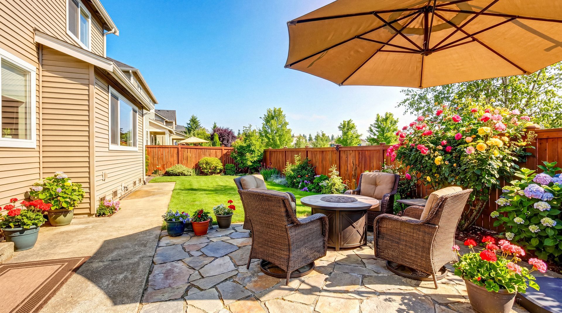 Sunlit backyard patio with wicker chairs, table, umbrella, flowers, and stone pavers beside a house