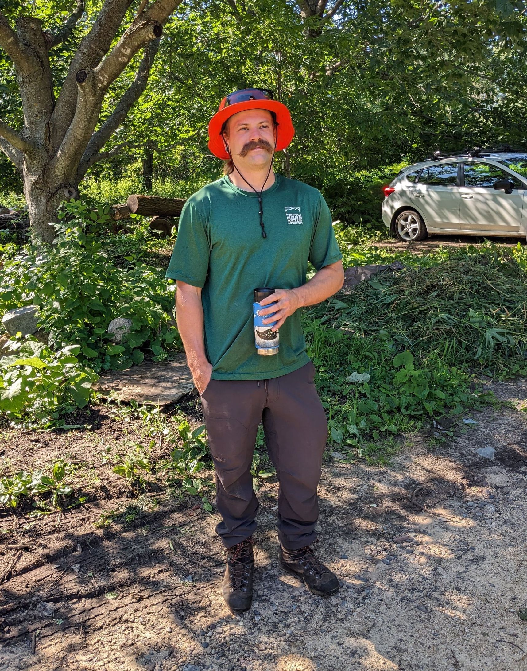 A man in a natural setting with an orange hat and big mustache.