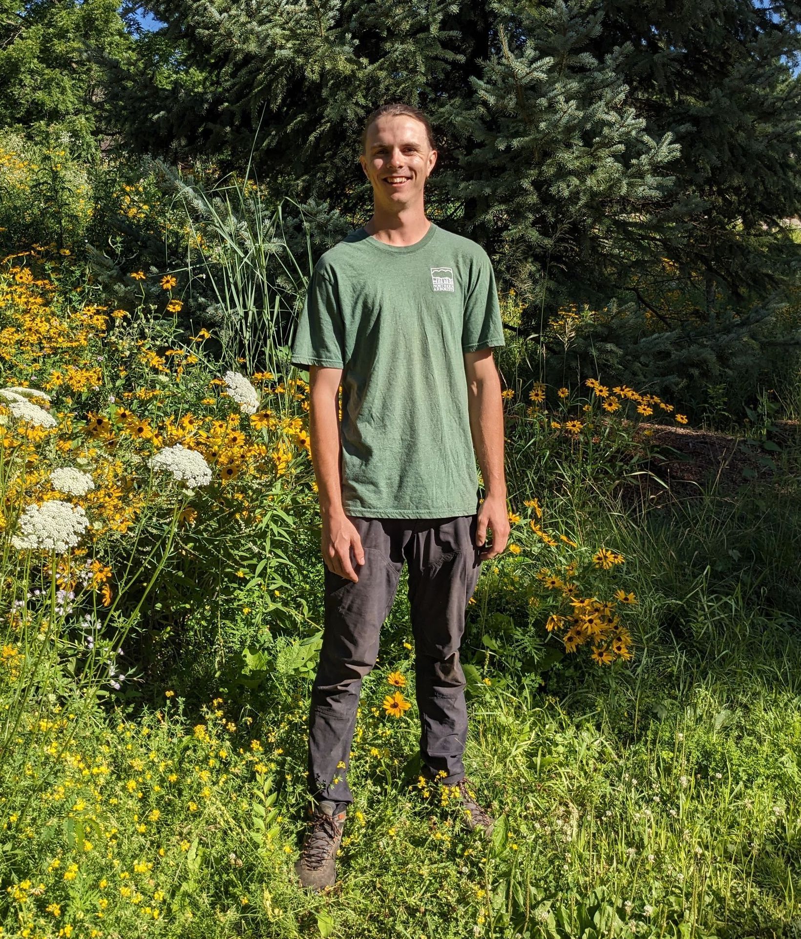 A tall man with a green shirt stands in some grass with prairie flowers behind.