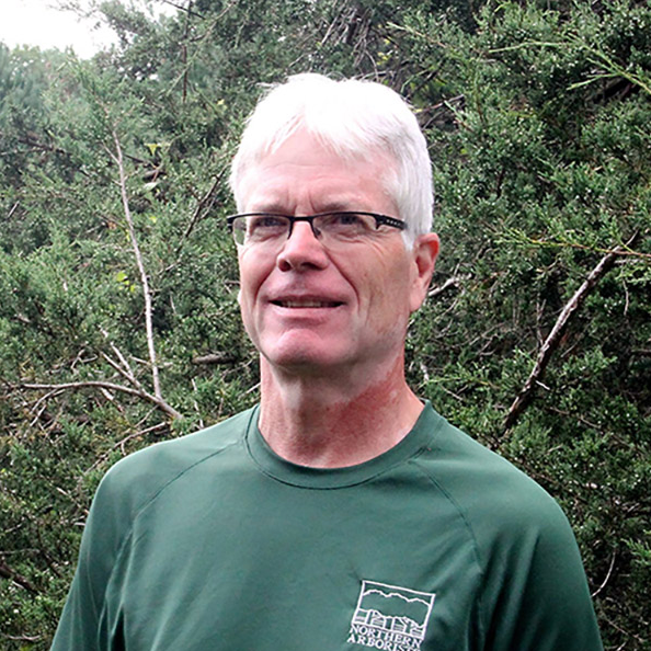 A man wearing glasses and a green shirt stands in front of trees