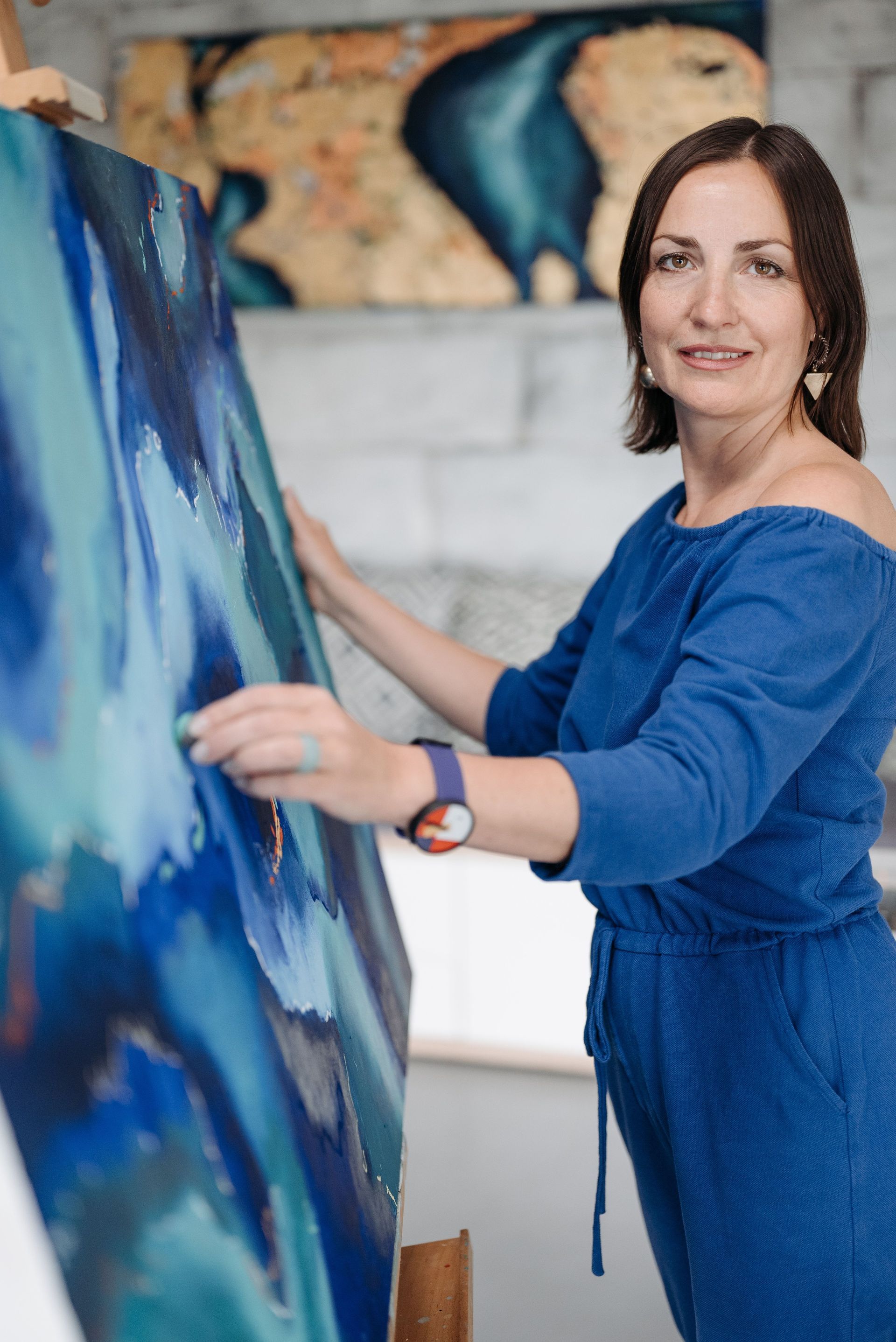 Vrouw in een blauwe jurk schildert op een groot doek in een lichte studio.