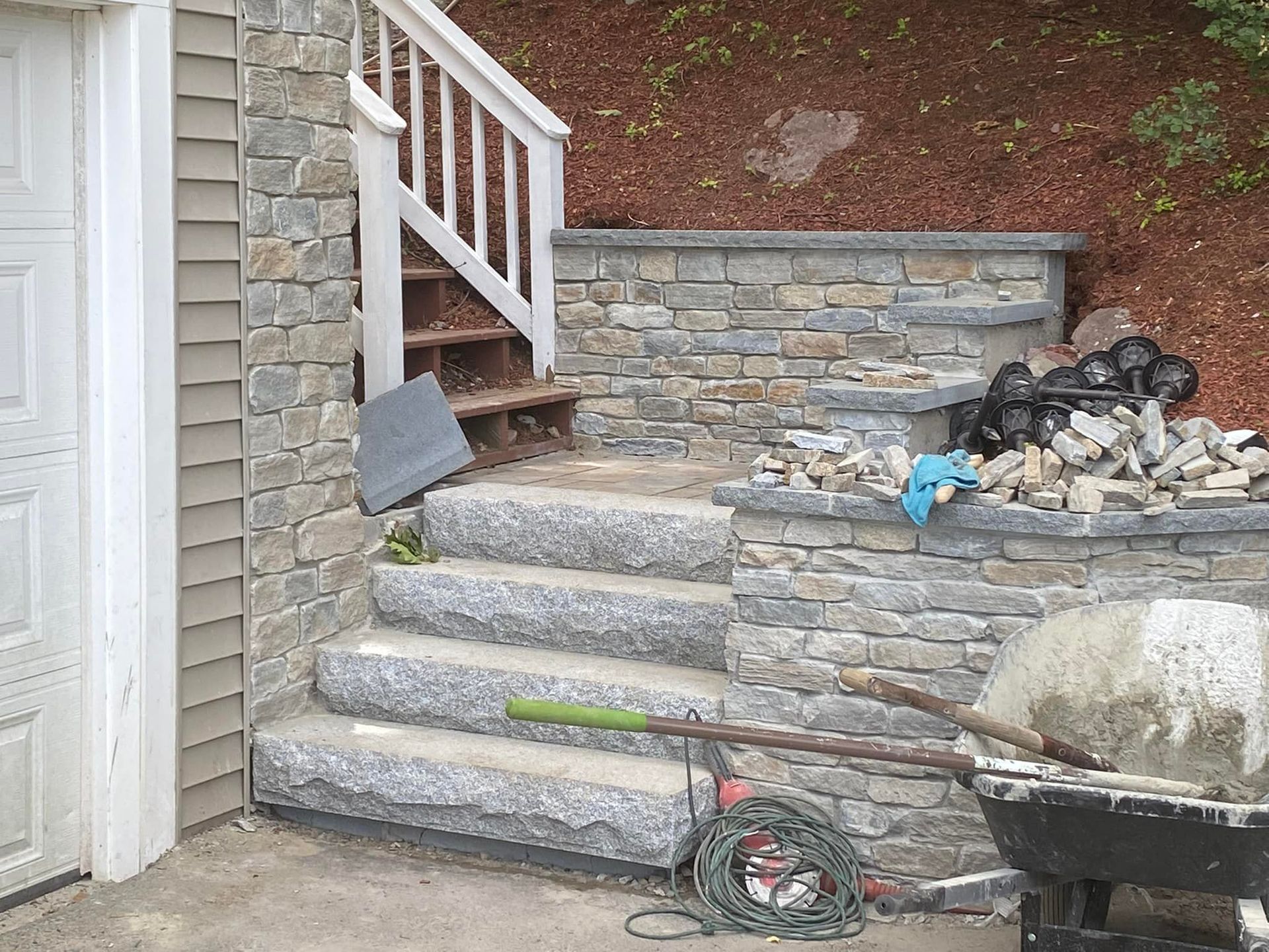 Stone steps and retaining wall under construction next to a garage; a wheelbarrow of concrete and tools are present.