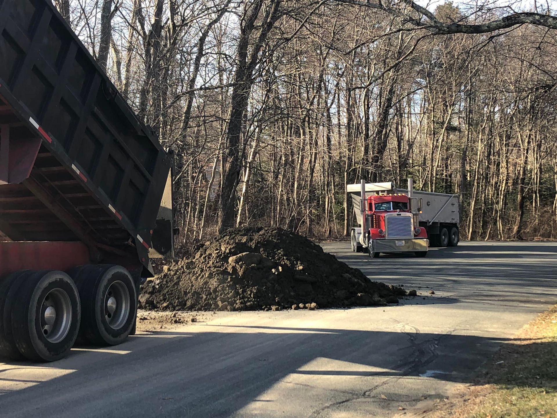 Dump truck unloading dirt onto a road, another truck in background near trees.